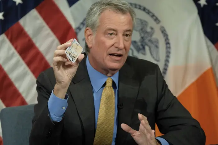 Mayor Bill de Blasio wearing a suit holds up an IDNYC card while sitting in front of flags at City Hall.